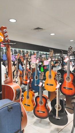 A music store display featuring various acoustic guitars hanging on racks. The guitars range in color, including natural wood tones, black, and blue. Several price tags are attached to the guitars, indicating a sale. The background reflects the inside of a music shop with bright lighting and mirrors enhancing the display.
