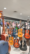 A music store display featuring various acoustic guitars hanging on racks. The guitars range in color, including natural wood tones, black, and blue. Several price tags are attached to the guitars, indicating a sale. The background reflects the inside of a music shop with bright lighting and mirrors enhancing the display.