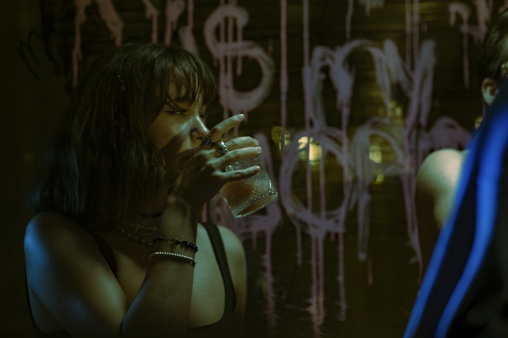 a woman drinking from a glass in front of a graffiti covered wall