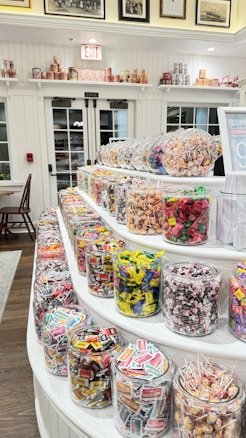Numerous jars filled with colorful assorted candies are displayed on a tiered white shelf. Various candy types include wrapped sweets and lollipops, offering a vibrant and inviting atmosphere. The background features white paneled walls and large windows with black doors, alongside shelves with additional tins and vintage photos.