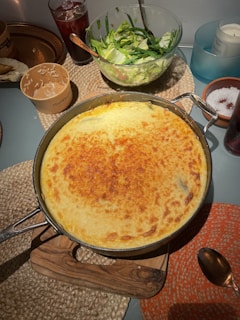 A baked dish with a golden, crispy top sits on a wooden board, surrounded by woven placemats. Next to it is a fresh green salad in a clear bowl, with lettuce and green beans visible. On the table, there are drinks, a small cup with a used spoon, and a dish with salt. The scene is set in a cozy dining environment.