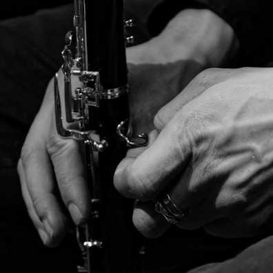 Hands holding a clarinet, focusing on the intricate details of the instrument's keys and the texture of the skin. The image is in black and white, highlighting shadows and contrasts.