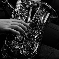 A close-up black and white photo of a person playing the saxophone, focusing on a hand pressing the keys. The instrument glistens with metallic reflections, showcasing its intricate design and craftsmanship.