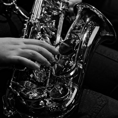 A close-up black and white photo of a person playing the saxophone, focusing on a hand pressing the keys. The instrument glistens with metallic reflections, showcasing its intricate design and craftsmanship.