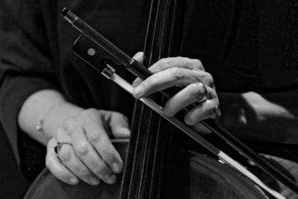A close-up black and white photograph of a person's hands playing a cello. The fingers of one hand are positioned on the strings and the other hand is holding a bow. The focus is on the hands and the instrument, capturing the texture and detail of both.