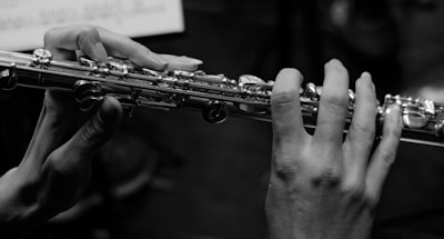 Hands are holding a flute, with fingers positioned on the keys. The image is in black and white, highlighting the details on the metallic surface of the flute. Music sheets are visible in the background, slightly out of focus.