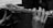 Hands are holding a flute, with fingers positioned on the keys. The image is in black and white, highlighting the details on the metallic surface of the flute. Music sheets are visible in the background, slightly out of focus.