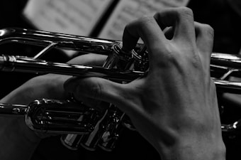 A black and white close-up of a person's hand playing a trumpet, focusing on the intricate details of the fingers pressing the trumpet valves. The background is softly out of focus, with sheet music visible but blurred.