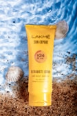 A tube of Lakmé Sun Expert Ultramatte Lotion with SPF 24 is placed on a textured surface that resembles a mixture of sand and water. The sunscreen is accompanied by seashells, creating a beach-like atmosphere. The package is bright yellow, making it stand out against the natural background.