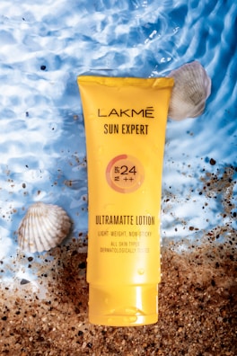 A tube of Lakmé Sun Expert Ultramatte Lotion with SPF 24 is placed on a textured surface that resembles a mixture of sand and water. The sunscreen is accompanied by seashells, creating a beach-like atmosphere. The package is bright yellow, making it stand out against the natural background.
