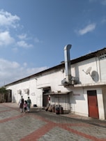 A long, single-story industrial building with a white exterior and a red door is in view. Several large industrial chimneys and satellite dishes are attached to the building. Two women and a child walk along the paved area of red and grey interlocking tiles. LPG gas cylinders and other equipment are stored along the side of the building. The sky is clear and blue with a few clouds.