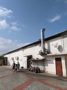 A long, single-story industrial building with a white exterior and a red door is in view. Several large industrial chimneys and satellite dishes are attached to the building. Two women and a child walk along the paved area of red and grey interlocking tiles. LPG gas cylinders and other equipment are stored along the side of the building. The sky is clear and blue with a few clouds.