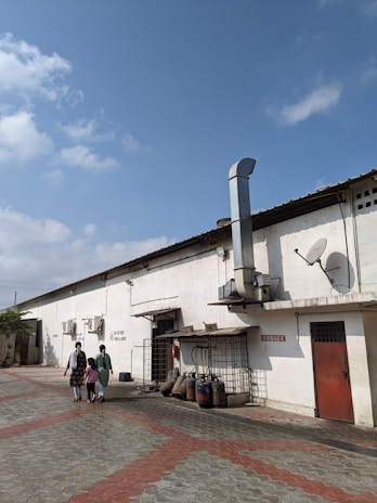 A long, single-story industrial building with a white exterior and a red door is in view. Several large industrial chimneys and satellite dishes are attached to the building. Two women and a child walk along the paved area of red and grey interlocking tiles. LPG gas cylinders and other equipment are stored along the side of the building. The sky is clear and blue with a few clouds.