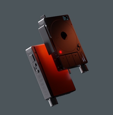 Two sleek, modern audio devices featuring a minimalist design with circular elements, buttons, and a display screen. The devices are primarily black with red accents and are positioned against a dark background.