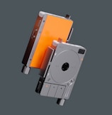 A pair of sleek, compact electronic devices are suspended in the air. They feature a minimalist design with a circular control interface on one side and a vibrant orange panel on the other. The devices have dials and buttons for interaction, suggesting functionality related to audio or media control.