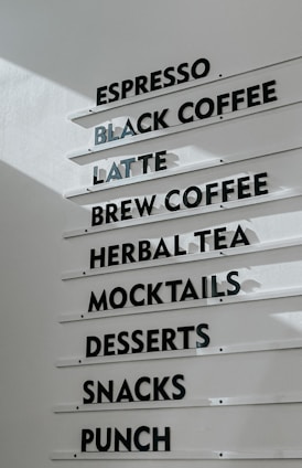 A menu board displaying various beverage and food options, including espresso, black coffee, latte, brew coffee, herbal tea, mocktails, desserts, snacks, and punch. The text is in black capital letters on white horizontal panels attached to a wall.