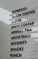 A menu board displaying various beverage and food options, including espresso, black coffee, latte, brew coffee, herbal tea, mocktails, desserts, snacks, and punch. The text is in black capital letters on white horizontal panels attached to a wall.
