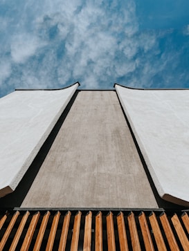 An upward perspective of a modern architectural structure with a minimalistic design. The building features smooth, light-colored concrete walls and a lower section with wooden slats. The sky above is clear with wispy clouds, creating a contrast between the solid structure and the soft sky.