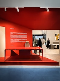 A modern office interior showcasing a sleek red desk with a matching chair, set against a vibrant red wall. Several people are visible in the background, interacting within the spacious office area. A large text block on the left side of the red wall provides information about the company.