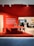 A modern office interior showcasing a sleek red desk with a matching chair, set against a vibrant red wall. Several people are visible in the background, interacting within the spacious office area. A large text block on the left side of the red wall provides information about the company.