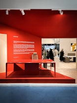 A modern office interior showcasing a sleek red desk with a matching chair, set against a vibrant red wall. Several people are visible in the background, interacting within the spacious office area. A large text block on the left side of the red wall provides information about the company.