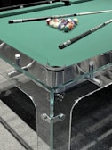 A pool table with a glass and metal frame, featuring a set of colorful pool balls arranged in a triangle formation at one end. Two pool cues rest on the green felt surface of the table.
