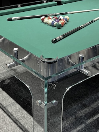 A pool table with a glass and metal frame, featuring a set of colorful pool balls arranged in a triangle formation at one end. Two pool cues rest on the green felt surface of the table.