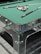 A pool table with a glass and metal frame, featuring a set of colorful pool balls arranged in a triangle formation at one end. Two pool cues rest on the green felt surface of the table.