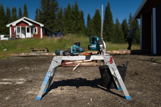 A workshop setting outside with a power saw, cordless drill, and other tools placed on a folding metal workbench. The background features a red and white house surrounded by grass and trees, adding a rural and peaceful atmosphere. A person is visible in the distance working on something near a structure. The sunlight casts a strong shadow of the tools on the ground.