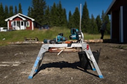 A workshop setting outside with a power saw, cordless drill, and other tools placed on a folding metal workbench. The background features a red and white house surrounded by grass and trees, adding a rural and peaceful atmosphere. A person is visible in the distance working on something near a structure. The sunlight casts a strong shadow of the tools on the ground.