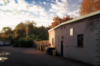 An industrial area featuring a long, light grey building with an outdoor storage area on the right. The building has small windows and various utility fixtures attached. The ground is paved, with puddles reflecting the surroundings. In the background, lush green trees and a few parked vehicles complete the scene under a sky dotted with fluffy clouds.