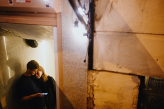 A couple stands closely together in a dimly lit, narrow space. The man is holding a phone, casting a blue light on their faces. The woman leans on his shoulder, creating an intimate atmosphere. The walls are textured, and the space feels cozy yet slightly enclosed.