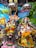 Two elaborately dressed deities are adorned with vibrant floral garlands, intricate jewelry, and colorful outfits. The background features a painted mural of nature with peacocks and trees, adding to the festive and sacred atmosphere.
