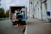 A person stands at the back of a moving truck on a city road, organizing a variety of items including boxes, a fan, and blue tarps. The truck is parked alongside a sidewalk bordered by a tall building with white stone walls and several business signs. The road appears empty except for parked cars and the scene is set under an overcast sky.