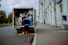 A person stands at the back of a moving truck on a city road, organizing a variety of items including boxes, a fan, and blue tarps. The truck is parked alongside a sidewalk bordered by a tall building with white stone walls and several business signs. The road appears empty except for parked cars and the scene is set under an overcast sky.