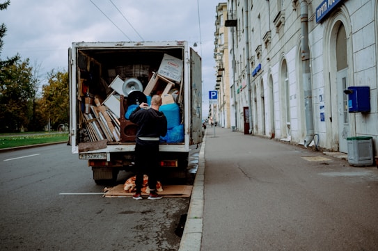 A person stands at the back of a moving truck on a city road, organizing a variety of items including boxes, a fan, and blue tarps. The truck is parked alongside a sidewalk bordered by a tall building with white stone walls and several business signs. The road appears empty except for parked cars and the scene is set under an overcast sky.