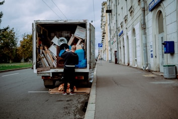 A person stands at the back of a moving truck on a city road, organizing a variety of items including boxes, a fan, and blue tarps. The truck is parked alongside a sidewalk bordered by a tall building with white stone walls and several business signs. The road appears empty except for parked cars and the scene is set under an overcast sky.