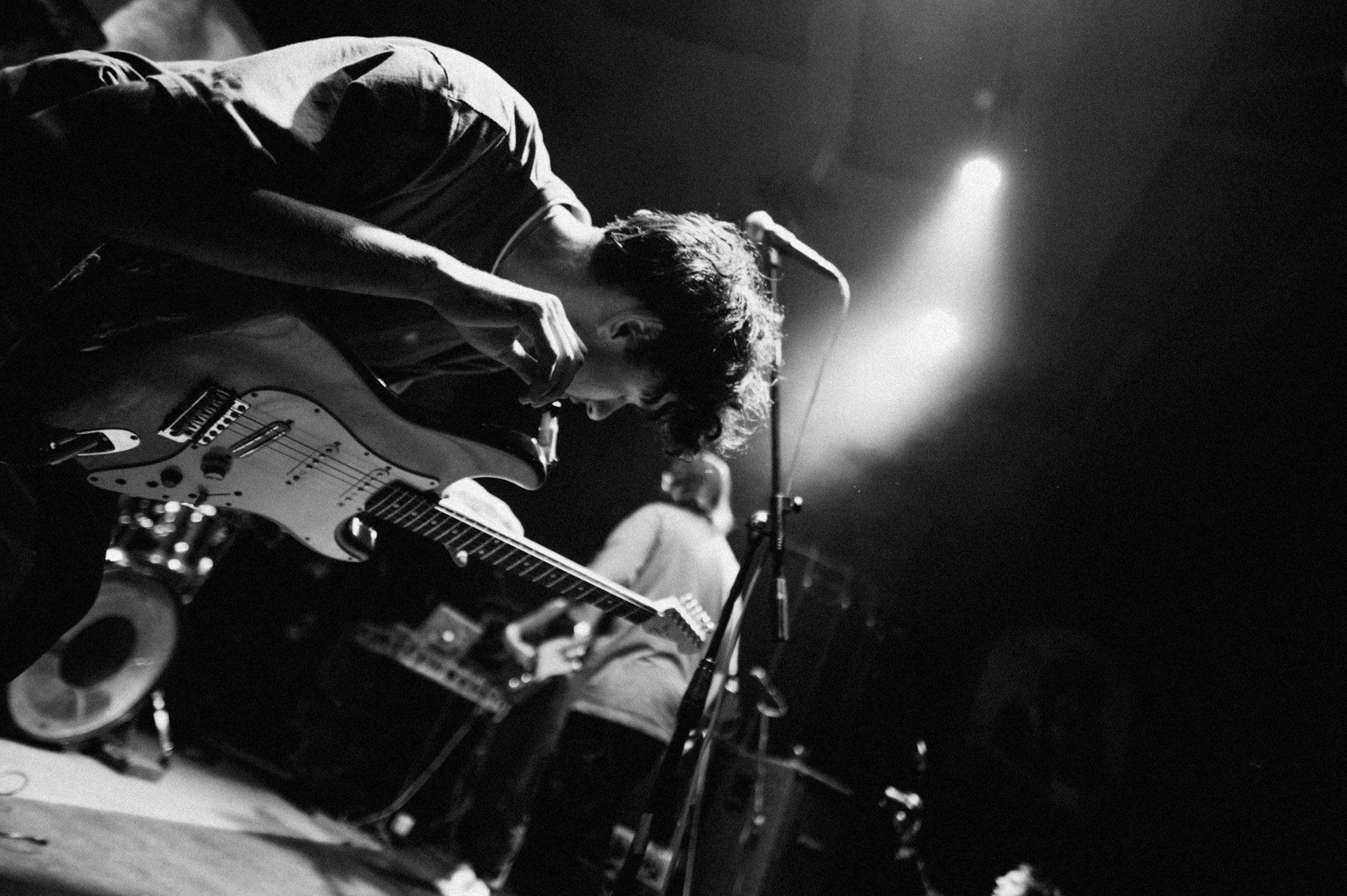 A black and white photograph of a musician intensely playing an electric guitar on stage. The performer is bent over, absorbed in the music, with a microphone nearby. Stage lighting creates dramatic shadows and highlights. Additional equipment and another musician are partially visible in the background.