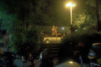 A group of people are playing volleyball under bright artificial lights in an outdoor setting at night. The surrounding area is dark, with trees and vegetation around the court. Motorcycles are parked in the foreground, and the entire scene has a subdued, night-time ambiance.