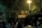A group of people are playing volleyball under bright artificial lights in an outdoor setting at night. The surrounding area is dark, with trees and vegetation around the court. Motorcycles are parked in the foreground, and the entire scene has a subdued, night-time ambiance.