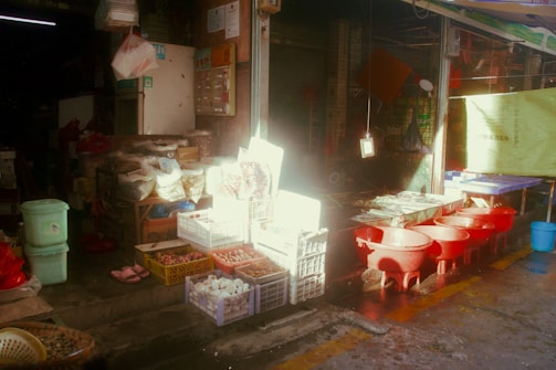 A market stall with various goods including baskets of eggs and other produce is displayed beside red basins. The setting appears to be indoors with soft, diffuse lighting creating a somewhat serene yet bustling atmosphere. Several items are stored in boxes and bags, and there is a small step leading into a darker area with shelves and more goods.
