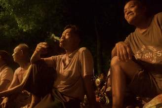 A group of men, primarily older, sit outdoors during the evening. One man is smoking, and the group appears relaxed and contemplative. The setting is likely a park or open area with trees in the background. The lighting is warm, giving a serene and nostalgic atmosphere.