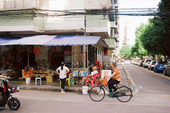 A street-side shop with various colorful items displayed under a blue awning. Several people are nearby, including a person seated and another standing. A person rides a bicycle past the shop, and a scooter can be seen partially in the frame. The street is lined with parked cars and trees, and the buildings have a worn, urban look.