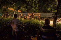 A group of people are gathered to watch and participate in a night volleyball game surrounded by lush greenery. The scene is dimly lit, with shadows cast by the lights illuminating the playing area. Some spectators are seated, while others stand by the net.