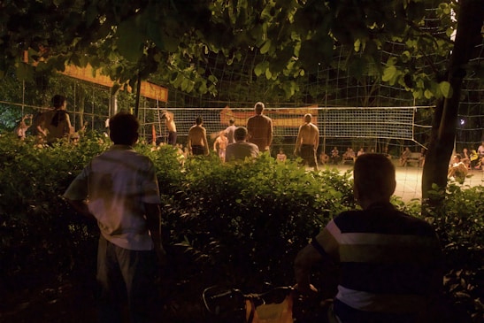 A group of people are gathered to watch and participate in a night volleyball game surrounded by lush greenery. The scene is dimly lit, with shadows cast by the lights illuminating the playing area. Some spectators are seated, while others stand by the net.