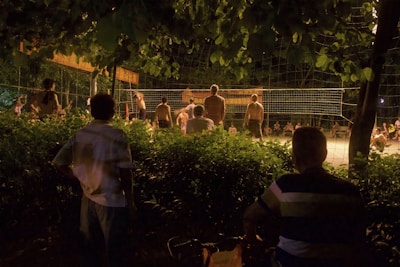 A group of people are gathered to watch and participate in a night volleyball game surrounded by lush greenery. The scene is dimly lit, with shadows cast by the lights illuminating the playing area. Some spectators are seated, while others stand by the net.