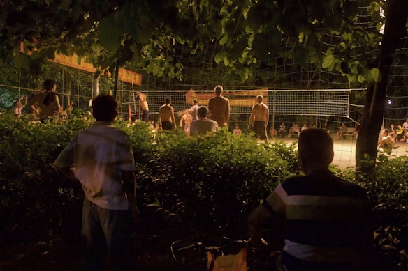 A group of people are gathered to watch and participate in a night volleyball game surrounded by lush greenery. The scene is dimly lit, with shadows cast by the lights illuminating the playing area. Some spectators are seated, while others stand by the net.