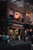 A bustling street scene in front of a brick building featuring a neon sign reading Savannah's Candy Kitchen. The scene includes a group of people gathered and walking by, with strings of lights visible above. The atmosphere is lively, with warm lights illuminating the shop's interior.