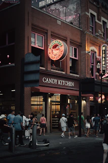 A bustling street scene in front of a brick building featuring a neon sign reading Savannah's Candy Kitchen. The scene includes a group of people gathered and walking by, with strings of lights visible above. The atmosphere is lively, with warm lights illuminating the shop's interior.