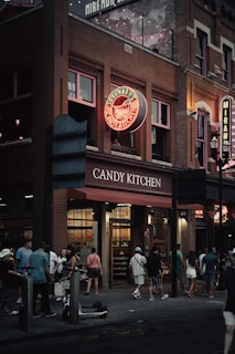 A bustling street scene in front of a brick building featuring a neon sign reading Savannah's Candy Kitchen. The scene includes a group of people gathered and walking by, with strings of lights visible above. The atmosphere is lively, with warm lights illuminating the shop's interior.